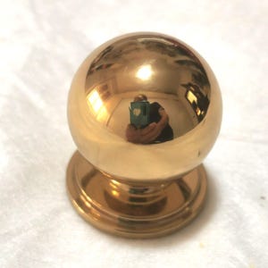 May include: A shiny, gold-colored cabinet knob with a spherical top and a circular base. The knob reflects the surrounding environment. This hardware is suitable for furniture restoration or new builds.