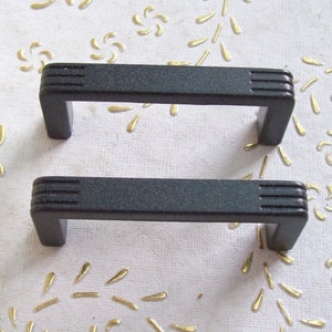 Peut inclure: Deux poignées de tiroir en plastique noir de forme rectangulaire et légèrement incurvée. Les poignées ont une surface texturée et mesurent environ 7,5 cm de long.