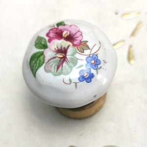 Könnte beinhalten: Weißer Keramik-Schrankknopf mit floralem Design mit rosa, lila und blauen Blumen mit grünen Blättern. Der Knopf ist auf einem kleinen, runden Holzsockel montiert. Dekorative Möbelbeschläge.