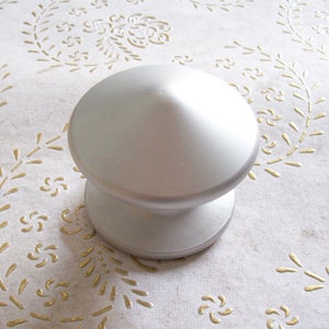 May include: A silver-colored, round, decorative knob with a pointed top. The knob is sitting on a white surface with a gold floral pattern.