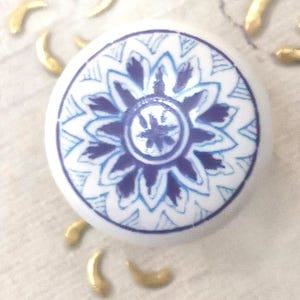 May include: A white ceramic knob with a blue floral design. The knob features a detailed pattern of blue petals and a central design. Gold-colored decorative accents surround the knob, creating a decorative aesthetic.