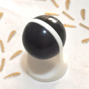 May include: A black and white bottle stopper with a spherical top and a white base. The black sphere has a white stripe around its center. The stopper is on a white surface with small, tan, seed-like objects scattered around it.