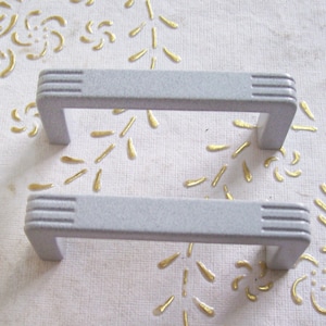 May include: Two white plastic drawer pulls with a rectangular shape and a raised design on the top. The pulls are on a white surface with a gold floral pattern.