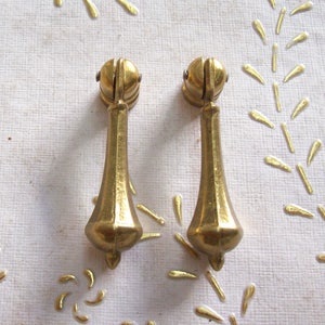May include: Two gold-toned metal cabinet pulls with a decorative, ornate design. The pulls have a rounded top and a pointed bottom.