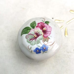 May include: A small, round, white ceramic knob featuring a floral design. The flowers are in shades of pink, green, and blue, with green leaves. The knob is likely for a cabinet or drawer, adding a touch of vintage charm.