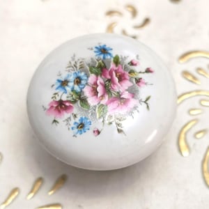Może przedstawiać: Biała, okrągła gałka ceramiczna z kwiatowym wzorem. Kwiaty są różowe i niebieskie, z zielonymi liśćmi. Gałka jest prawdopodobnie przeznaczona do szafki lub szuflady.