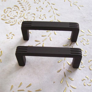 May include: Two black cabinet pulls with a rectangular shape and a slightly curved top. The pulls have a grooved design on the top and bottom.