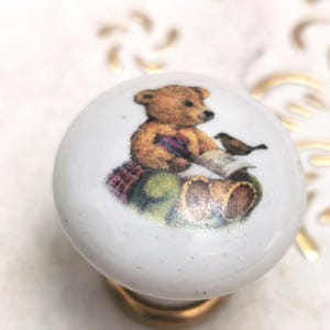 May include: White ceramic cabinet knob with a gold base. The knob features a detailed illustration of a teddy bear with a bird. The bear is wearing a scarf and sitting on a pillow. The knob is a decorative accent for furniture.
