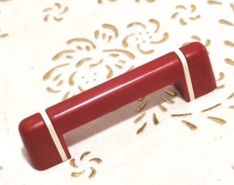 May include: A red cabinet pull with white accents. The rectangular handle is set against a white surface with gold floral patterns. This hardware is suitable for drawers and cabinets.