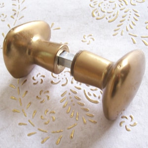 May include: Two gold-toned door knobs, one with a round shape and the other with an oval shape, are connected by a metal rod.