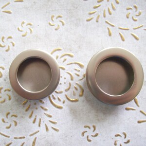 May include: Two silver-toned metal drawer pulls with a round, recessed centre. The pulls are on a white background with a gold floral pattern.