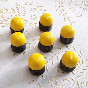 May include: Seven yellow and black round buttons with a smooth, glossy finish. The buttons are arranged in a semi-circle pattern on a white surface with a floral design.
