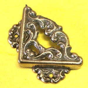 May include: A brass-colored, corner bracket with ornate, swirling scrollwork. The right-angled bracket has a decorative open center and small mounting holes. The background is a solid yellow.