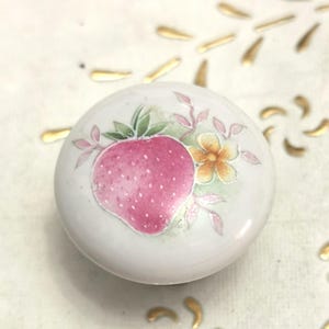 May include: A white ceramic knob with a hand-painted strawberry design. The strawberry is pink with white speckles, surrounded by green leaves, pink accents, and a small yellow flower. The knob is likely for a cabinet or drawer.