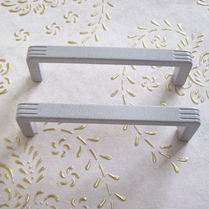 May include: Two gray cabinet pulls with a textured finish. The pulls are rectangular with rounded edges and have a simple, modern design.