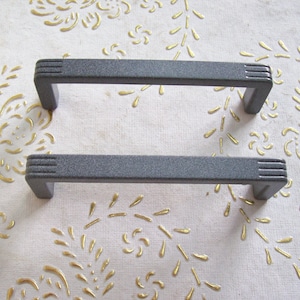 May include: Two grey metal cabinet door pulls with a textured finish. The pulls have a rectangular shape with rounded edges.