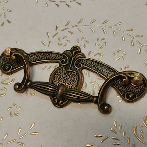 Peut inclure: Une poignée de tiroir en laiton antique décorative avec un design orné. La poignée a une forme incurvée avec une poignée centrale et deux extrémités décoratives.