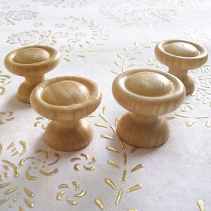 May include: Four wooden knobs with a round top and a cylindrical base. The knobs are a light brown color and are arranged in a semi-circle on a white surface with a gold pattern.