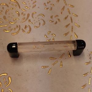 Peut inclure: Une poignée de tiroir en plastique transparent avec des embouts noirs. La poignée est montée sur une surface blanche avec un motif floral doré.