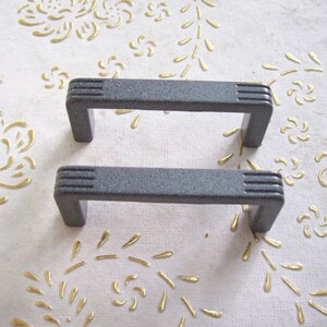 May include: Two grey metal cabinet door pulls with a textured finish. The pulls are rectangular with rounded edges and have a square shape in the centre.