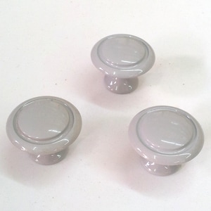 Peut inclure: Trois boutons de cabinet en céramique gris avec un bord surélevé. Les boutons ont un dessus rond et une base cylindrique courte.