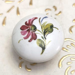 Puede incluir: Pomo de cerámica blanca con un diseño floral pintado a mano. La flor es de color rojo intenso con hojas verdes y amarillas. El pomo es redondo y tiene una superficie lisa. Herrajes decorativos.