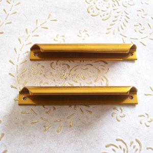 Puede incluir: Dos tiradores de cajón de metal de color dorado, con forma rectangular y un corte curvo en el centro.