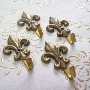 Puede incluir: Cuatro ganchos de metal dorado en forma de flor de lis. Los ganchos están montados en una superficie blanca.