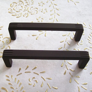 May include: Two brown metal cabinet door pulls with a rectangular shape and a slightly curved design.