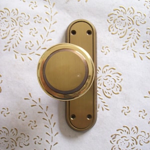 Op de afbeelding: Een gouden deurknop met een ronde knop en een rechthoekige plaat. De knop heeft een verhoogde ring rond de rand.