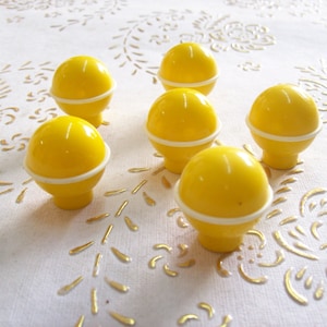 May include: Six yellow plastic knobs with white trim. The knobs are round and have a small stem on the bottom.