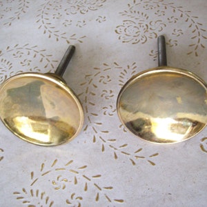 May include: Two gold-toned metal knobs with a round, slightly domed top and a long, thin metal stem. The knobs are on a white surface with a floral pattern.