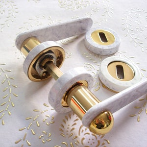 Peut inclure: Ensemble de poignées de porte en marbre blanc avec des accents dorés. L'ensemble comprend une poignée à levier, un bouton rond et deux rosaces.