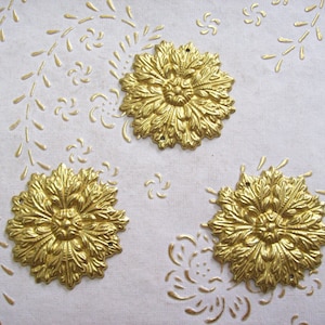 May include: Three gold-colored metal decorative embellishments with a floral design. The embellishments are on a white background with a subtle embossed pattern.