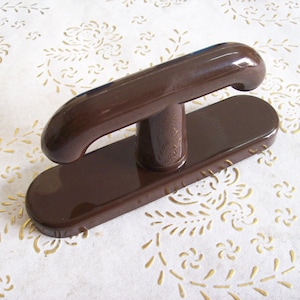 Op de afbeelding: Een bruine plastic deurgreep met een T-vormige greep. De greep is gemonteerd op een rechthoekige basis.