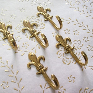 May include: Five gold-toned metal hooks with a fleur-de-lis design. The hooks are all in the same style and are arranged in a semi-circle on a white background.