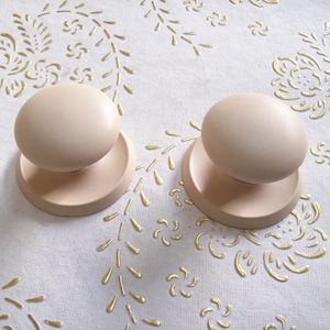 Puede incluir: Dos pomos de puerta de color beige claro con forma redonda y base plana. Los pomos están sobre una superficie blanca con un patrón floral dorado.