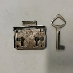 Puede incluir: Una cerradura de metal plateada con dos agujeros de llave y una llave a juego. La cerradura está ligeramente desgastada y tiene un aspecto rústico.