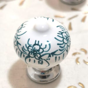 May include: A white ceramic cabinet knob with a floral design in teal. The knob has a rounded top and a silver-colored base. Decorative hardware for furniture.