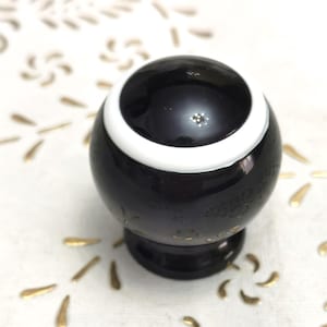 Könnte beinhalten: Ein glänzender, schwarzer Zierknauf mit einem weißen Ring am oberen Rand. Der Knauf steht auf einer weißen Oberfläche mit goldenen Blumenmustern. Wahrscheinlich für einen Schrank oder eine Schublade.