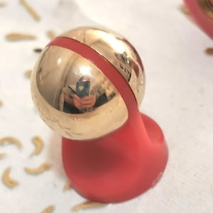 May include: A gold-colored sphere with a red band and a red base. The sphere reflects the surroundings. The base has a curved, organic shape. The object is on a white surface with small, tan-colored shapes scattered around it.