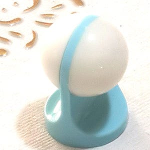 May include: A small, light blue and white decorative object. The object has a rounded white top with a light blue band and a light blue base. The base is a rounded shape. The object is sitting on a white surface.