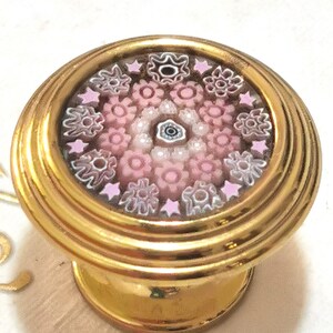 Può includere: Pomello per armadio dorato con un motivo floreale decorativo. Il pomello presenta una faccia circolare con un mosaico di fiori rosa e bianchi, circondata da un bordo dorato. La base è anch'essa dorata, con un design scanalato.