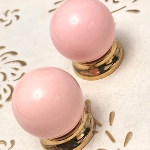 May include: Two pink, spherical cabinet knobs with a glossy finish. Each knob is mounted on a gold-coloured base with decorative accents. The knobs are set against a white background with gold leaf-like patterns.