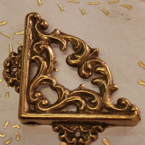 May include: A gold-colored metal corner bracket with a decorative, ornate design. The bracket has a scrolled, floral pattern.