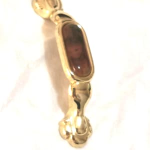 Op de afbeelding: Een goudkleurige kastgreep met een ovaal, bruin getint middenstuk. De greep heeft een decoratief ontwerp met een bloemachtig bovendeel en een klauwvoet. De hardware is waarschijnlijk voor meubels of kasten.