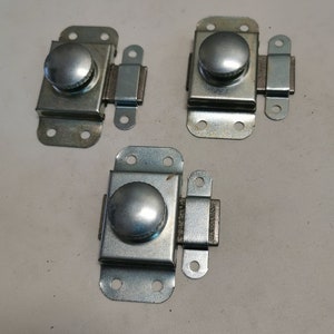 Op de afbeelding: Drie zilveren metalen kastdeurgrepen met een ronde knop. Elke greep heeft een rechthoekige plaat met vier gaten en een klein metalen greepje dat over de plaat schuift.