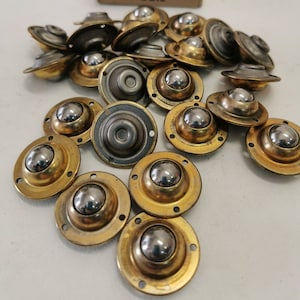 Puede incluir: Un conjunto de 18 ruedas de bolas de metal dorado con bolas de plata. Las ruedas están dispuestas en una pila sobre una superficie blanca. La caja de las ruedas es visible en el fondo. La caja es marrón con texto blanco que dice "Art 5008 mm. 30 6 Satz".