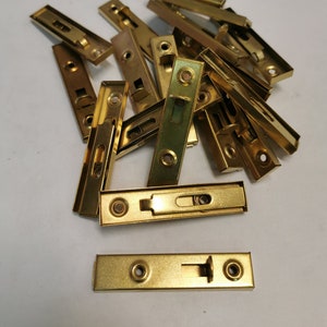 Könnte beinhalten: Ein Stapel goldfarbener Metall-Schrankscharniere. Die Scharniere sind rechteckig mit einem kleinen Riegel an einer Seite. Es befinden sich 15 Scharniere im Stapel.