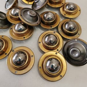 Puede incluir: Un conjunto de 16 ruedas de muebles de metal dorado con rodamientos de bolas. Cada rueda tiene una base redonda con un pequeño rodamiento de bolas en el centro. Las ruedas están dispuestas de forma aleatoria sobre una superficie blanca.